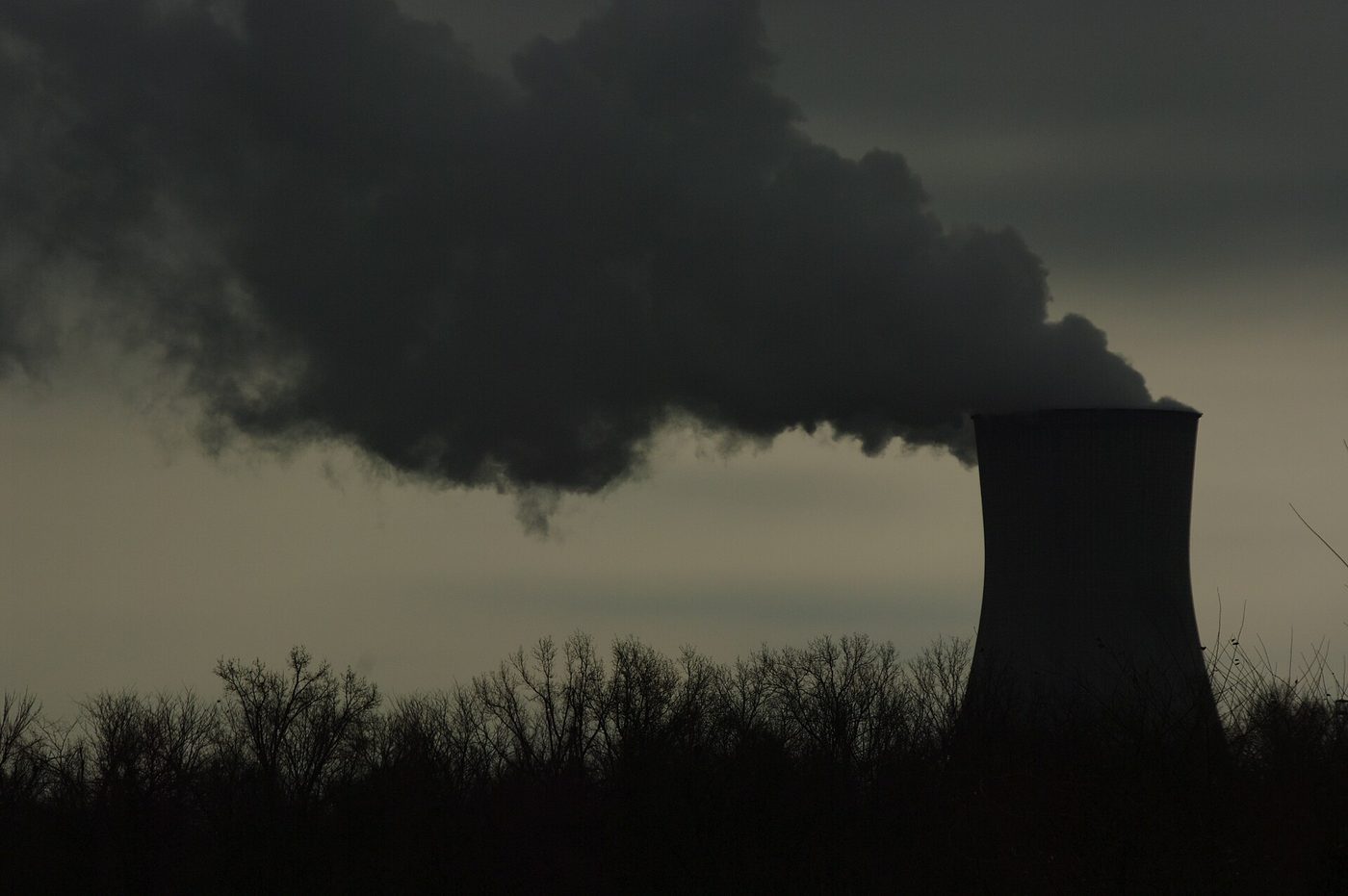 Photograph of the cooling tower of the Callaway nuclear power plant in Missouri in silhouette at dusk, releasing a large plume of water vapor into a dark, cloudy sky above a line of bare trees.