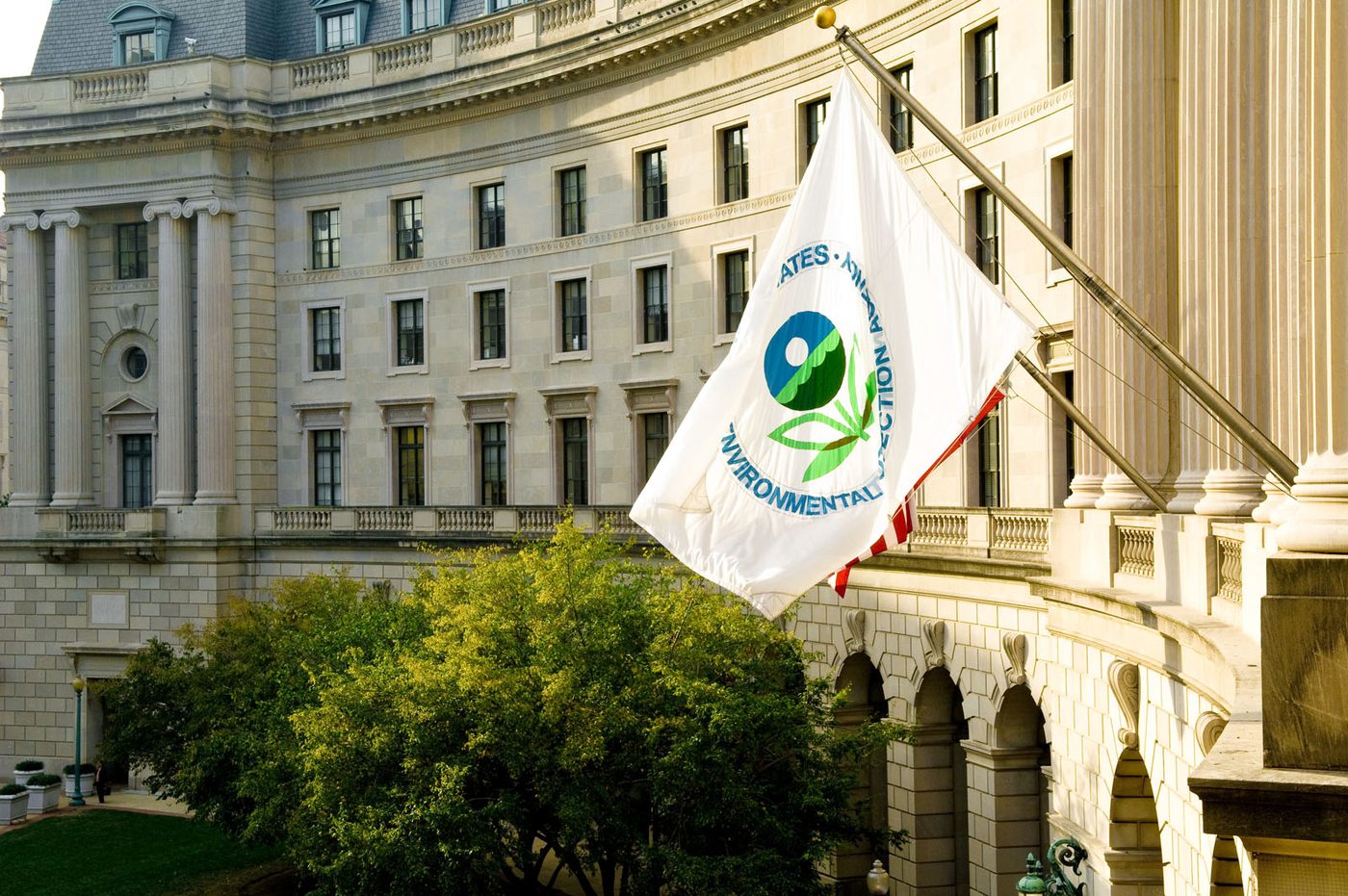 Photograph of the U.S. Environmental Protection Agency flag flying from a pole in front of the curved neoclassical stone facade of the William Jefferson Clinton Federal Building in Washington, D.C., with trees below.