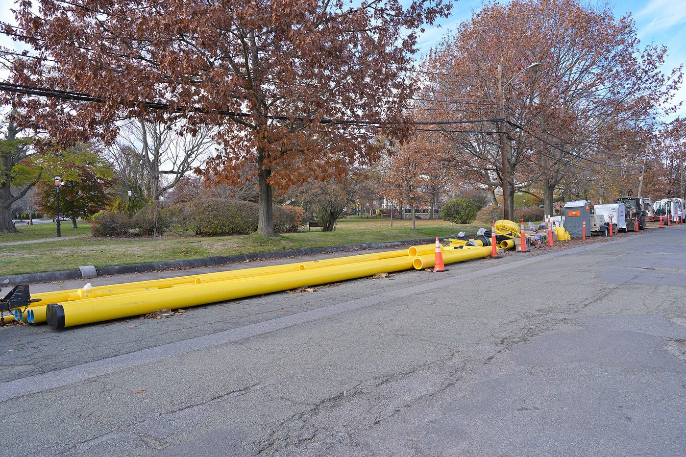Photograph of long yellow plastic natural gas pipes laid along the edge of a residential street in Newton, Massachusetts, staged for a gas main replacement project, with houses and parked cars in the background.