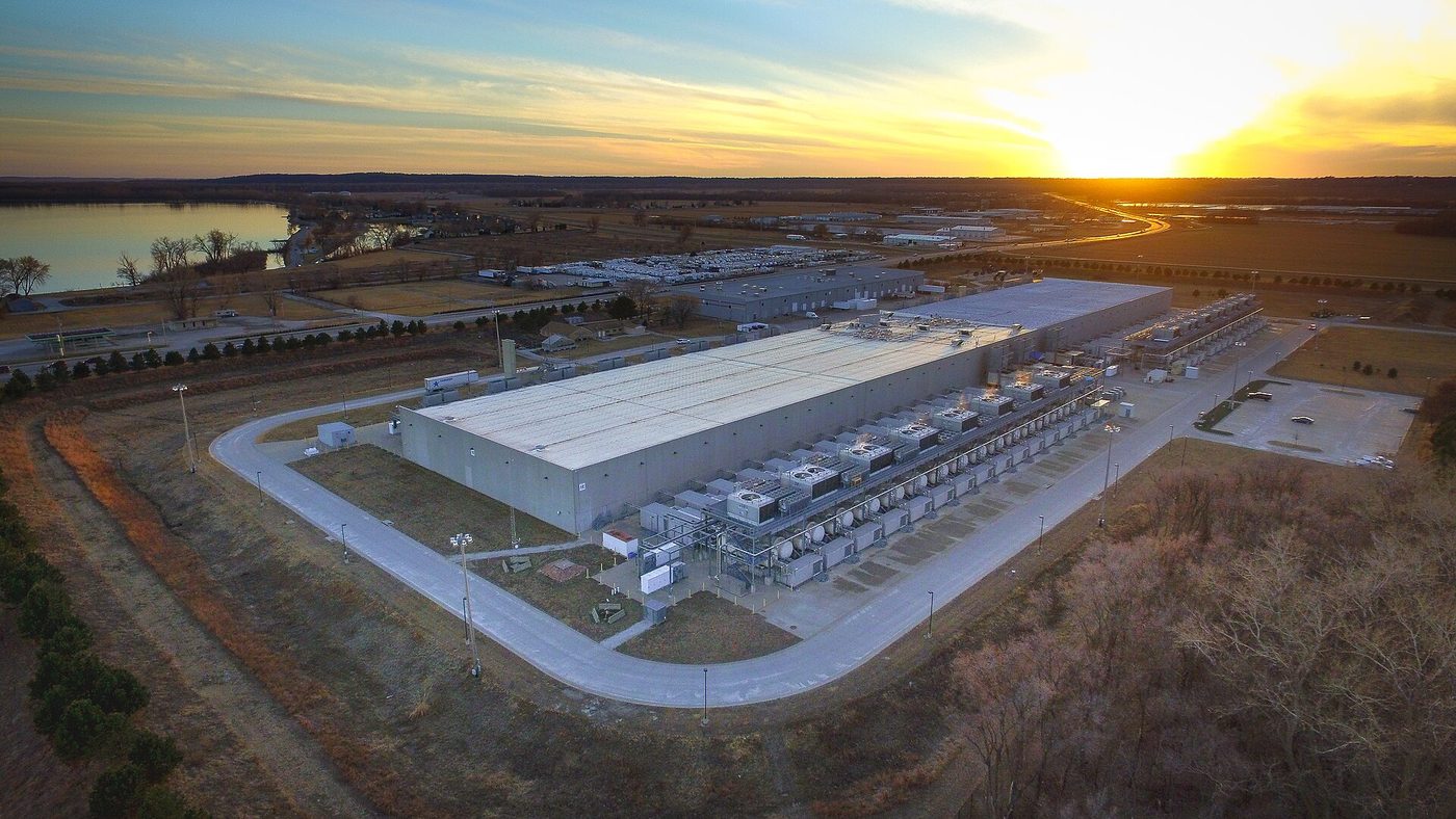 Aerial photograph at sunset of a Google data center in Council Bluffs, Iowa: a long, low, light-gray building with rows of rooftop and ground-level cooling equipment, surrounded by roads, fields and a river.