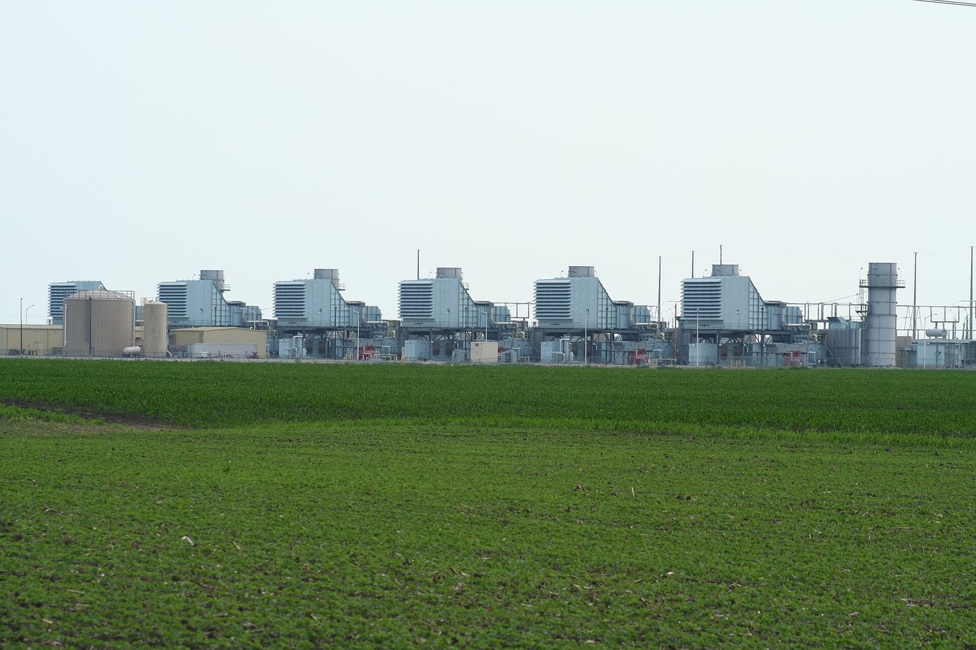 Photograph of the Goose Creek Energy Center in Piatt County, Illinois: a row of six natural gas combustion turbine units with boxy gray enclosures and short stacks, seen across a green crop field under a pale sky.