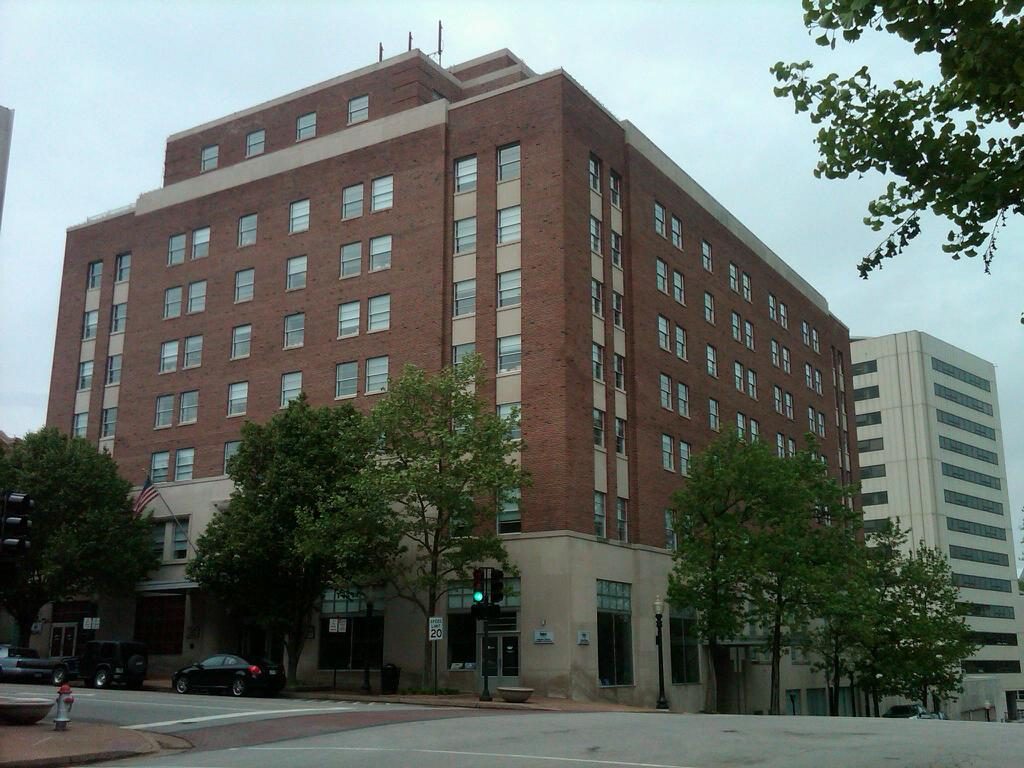 Photograph of the Governor Office Building in Jefferson City, Missouri, a tall red-brick office building with a light stone base on a street corner, with trees in front.