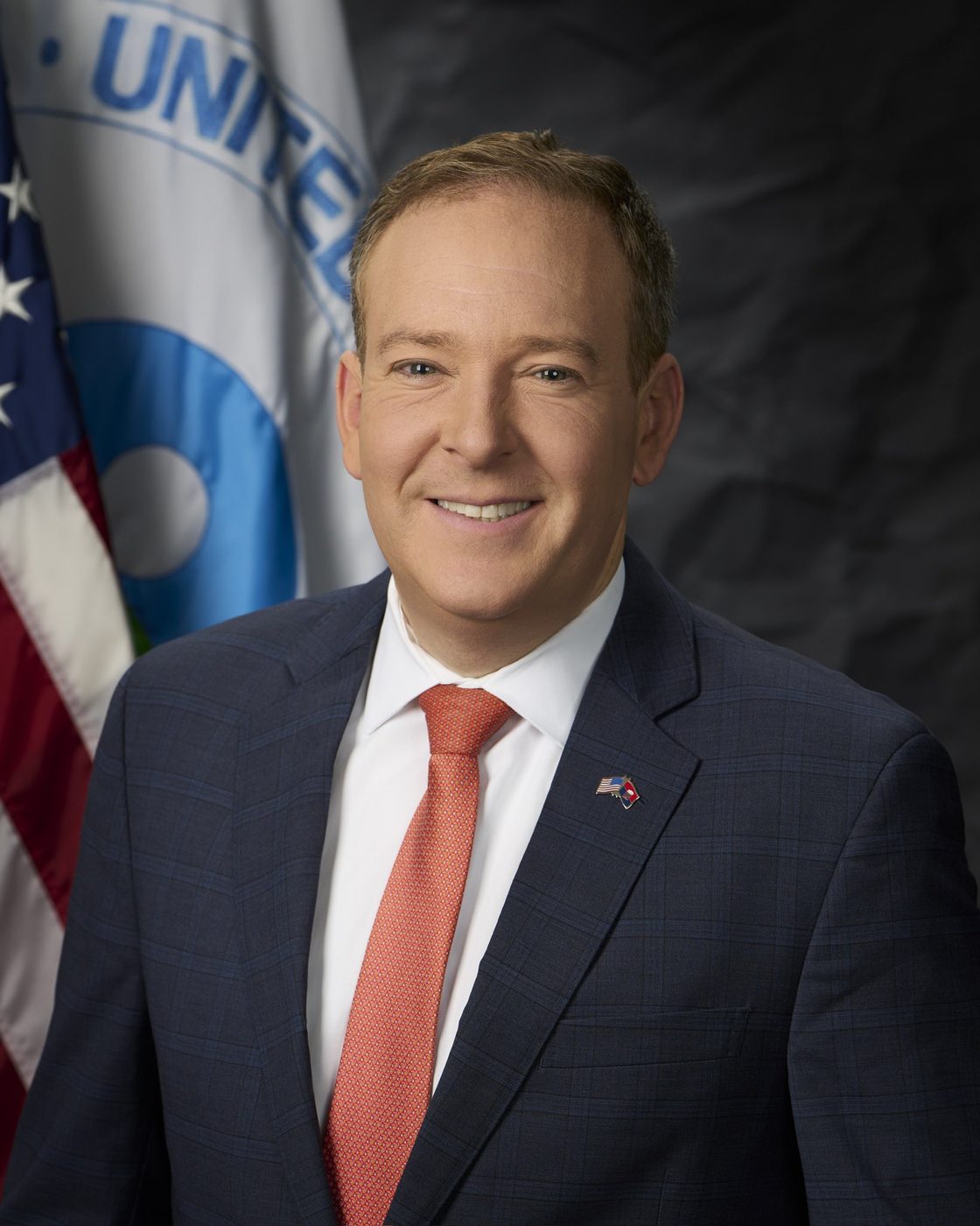 Official portrait photograph of EPA Administrator Lee Zeldin, smiling, in a dark blue suit, white shirt and coral tie, in front of the U.S. flag and the EPA flag.