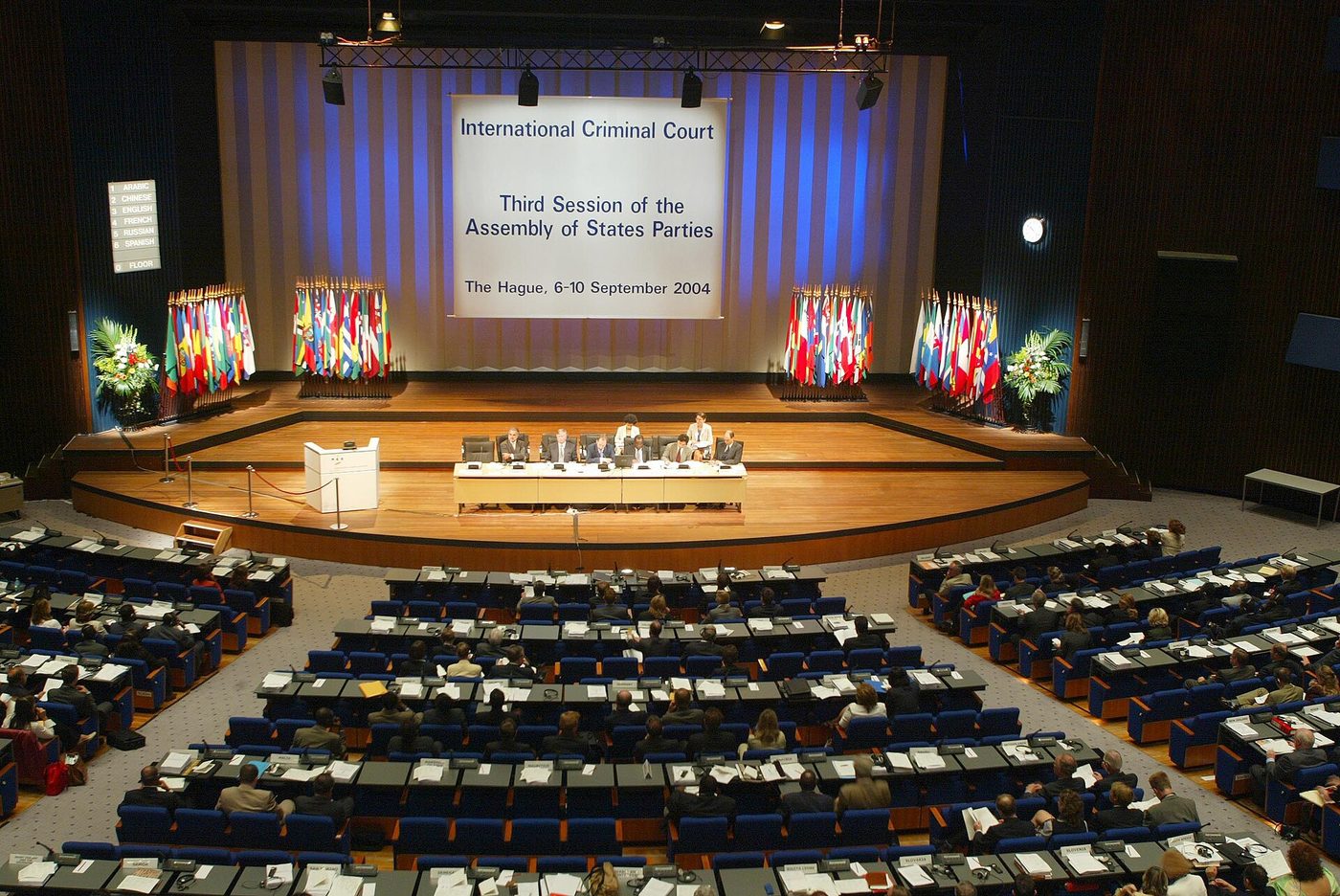 Photograph of a plenary session of the ICC Assembly of States Parties, with delegates at rows of desks facing a stage with national flags and a banner reading “International Criminal Court, Third Session of the Assembly of States Parties, The Hague, 6–10 September 2004.”