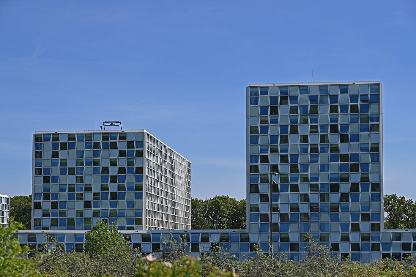 Photograph of the International Criminal Court’s permanent premises in The Hague, two towers with blue, green and grey glass panels rising behind trees and shrubs, seen from the Maurits Kiekpad path in May 2026.