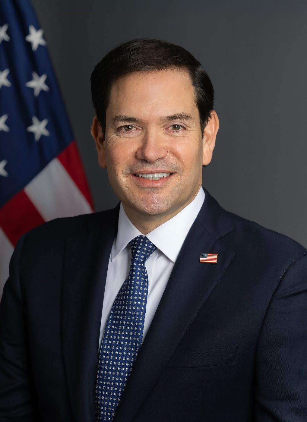 Official portrait photograph of Secretary of State Marco Rubio in a dark suit and patterned blue tie, smiling, beside the United States flag against a grey backdrop, January 2025.