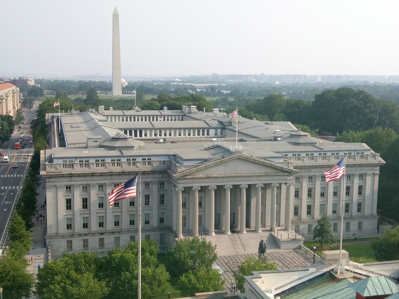 Photograph of the U.S. Treasury Building in Washington, D.C., seen from above: a large neoclassical stone building with a columned portico and U.S. flags, with the Washington Monument in the distance.